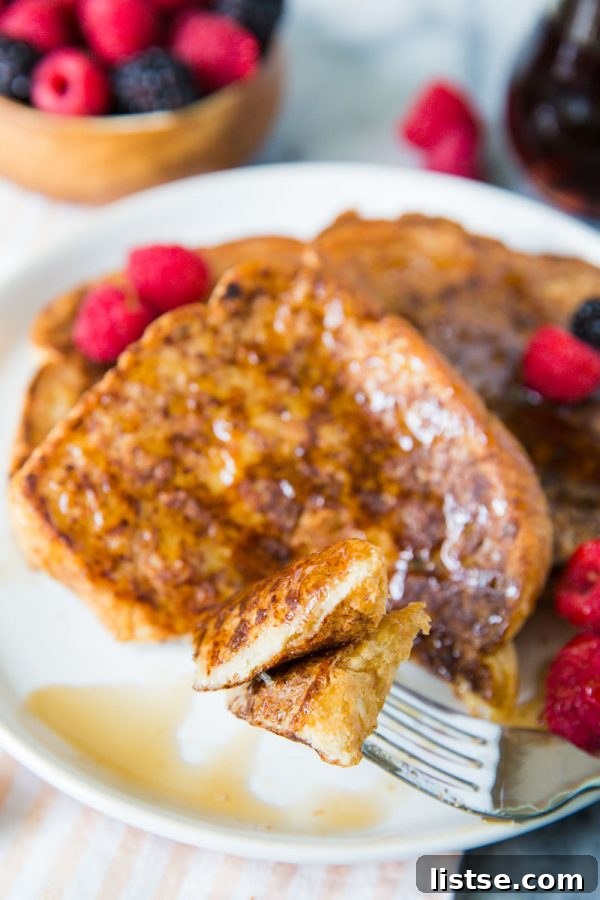 A close-up shot focusing on a fork with a perfect bite of vegan French toast, glistening with maple syrup and garnished with fresh berries, on a white plate with more slices visible in the background.