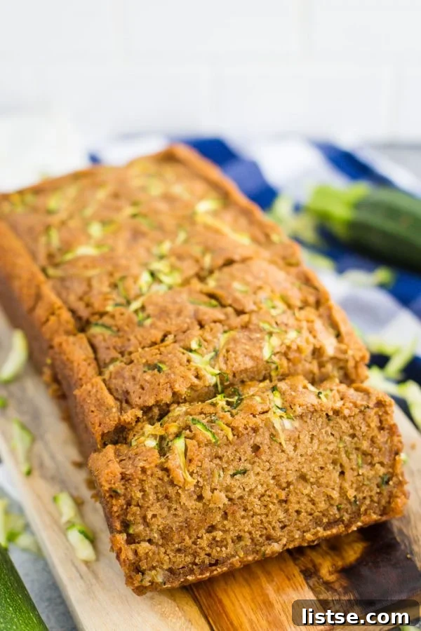 Moist Vegan Zucchini Loaf 2 A delectable loaf of vegan zucchini bread, freshly baked and sliced, resting elegantly on a rustic wooden board. In the soft-focus background, a charming blue checkered towel and a whole zucchini hint at the natural, wholesome ingredients used in this delightful summer recipe.