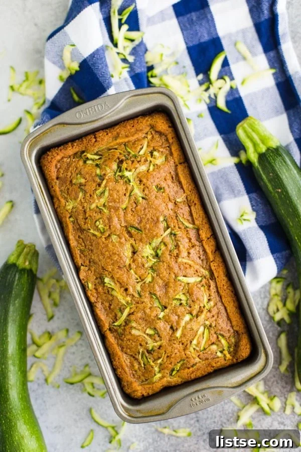 Moist Vegan Zucchini Loaf 4 A charming top-down perspective of freshly baked vegan zucchini bread nestled in a loaf pan, artfully placed on a blue checkered towel. Surrounding the pan are vibrant, fresh zucchinis, highlighting the star ingredient of this homemade delight.