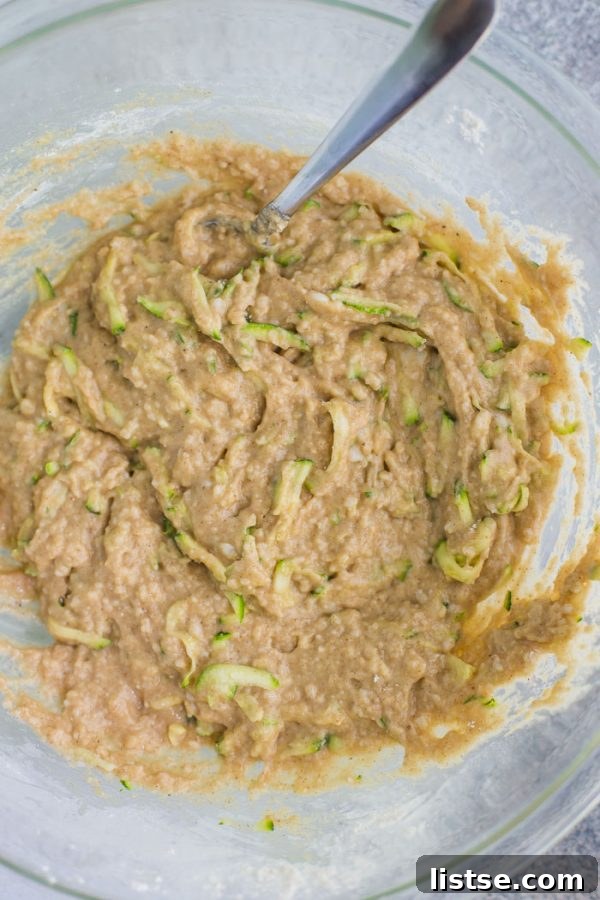 Moist Vegan Zucchini Loaf 6 A large glass bowl brimming with a rich, tan zucchini bread batter, prominently featuring generous chunks of shredded zucchini. The texture appears thick and ready for baking, promising a moist and flavorful loaf.