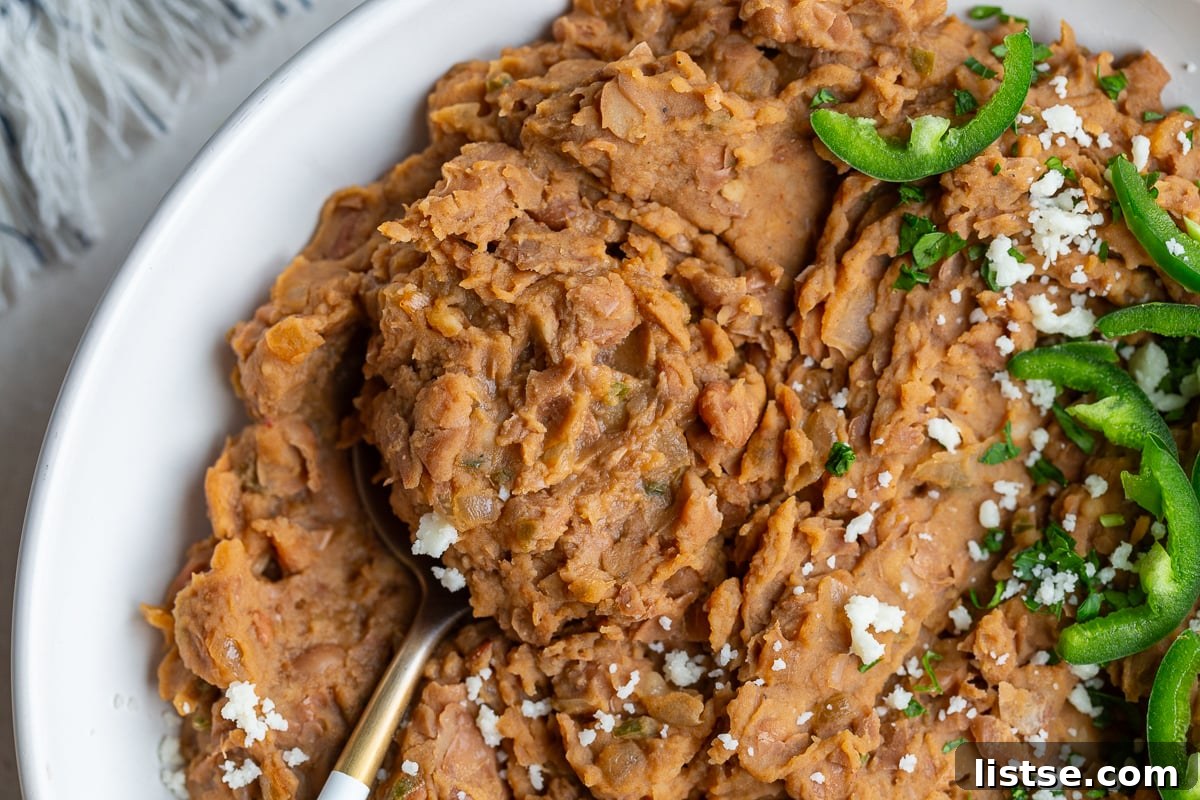 A close-up shot of creamy homemade refried beans served in a white bowl, garnished with a few fresh jalapeño slices and a spoon resting on the side