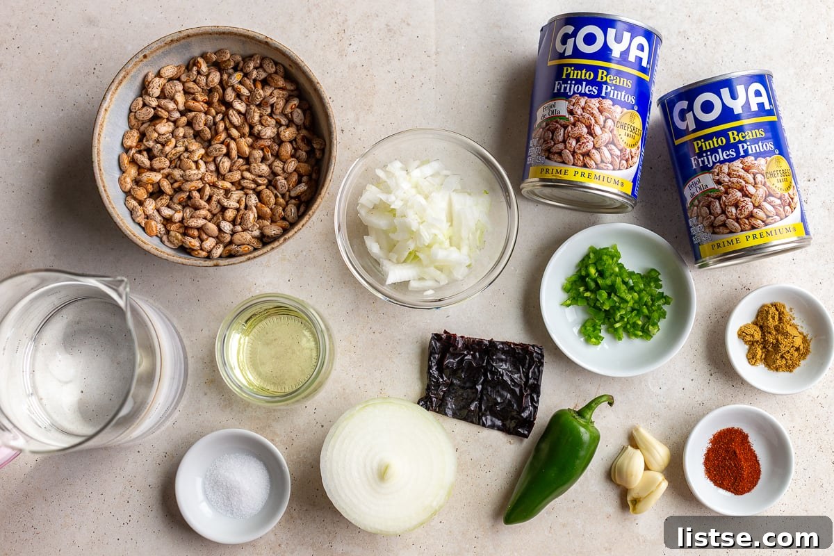 An organized display of fresh ingredients including dried pinto beans, a white onion, jalapeño, garlic cloves, and a selection of spices, set up for making refried beans from scratch