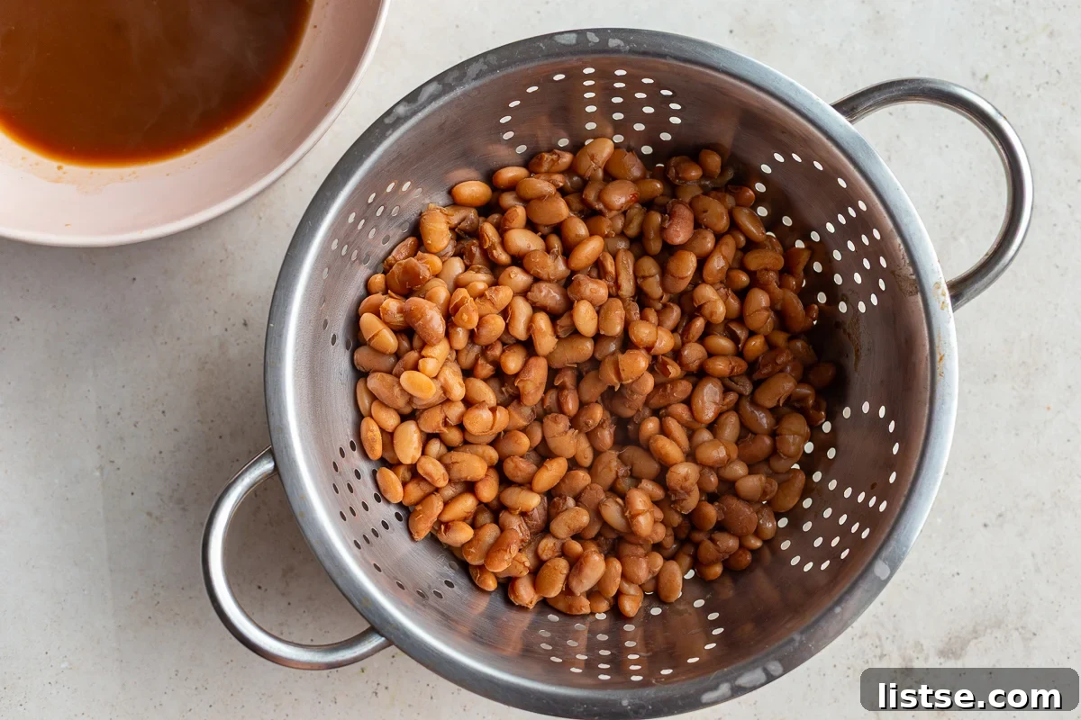 Cooked pinto beans draining in a metal colander, positioned over a bowl to collect the reserved bean cooking liquid