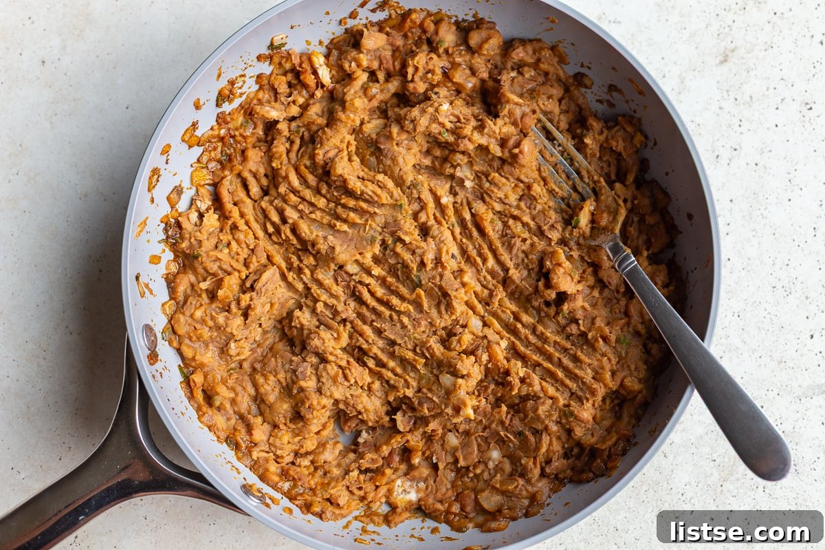 Cooked pinto beans being added to the sautéed aromatics in a skillet, preparing for the mashing process to create creamy refried beans