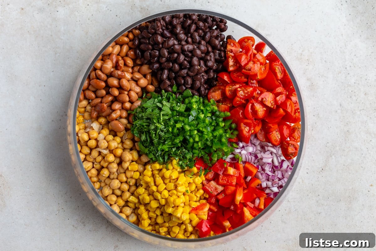 Bowl filled with neatly sectioned ingredients for Southwest bean salad including pinto beans, black beans, chickpeas, tomato, onion, jalapeno, corn and bell pepper