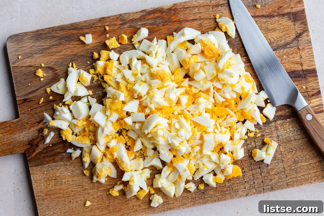 Finely chopped hard-boiled eggs, including both whites and creamy yolks, are neatly arranged on a rustic wooden cutting board, ready to be mixed into a flavorful egg salad.