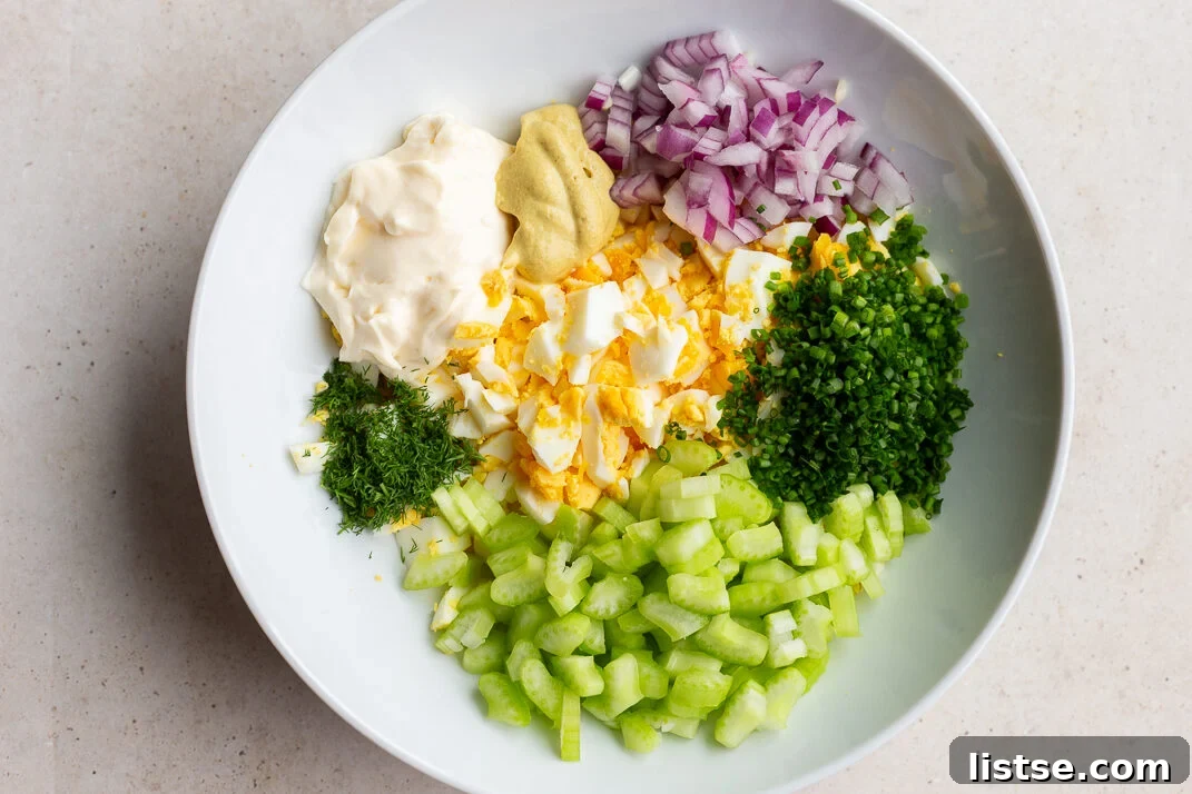 A large mixing bowl filled with the vibrant ingredients for homemade egg salad, including chopped hard-boiled eggs, diced red onion, fresh chives, creamy mayonnaise, and crisp celery, awaiting a thorough mix.