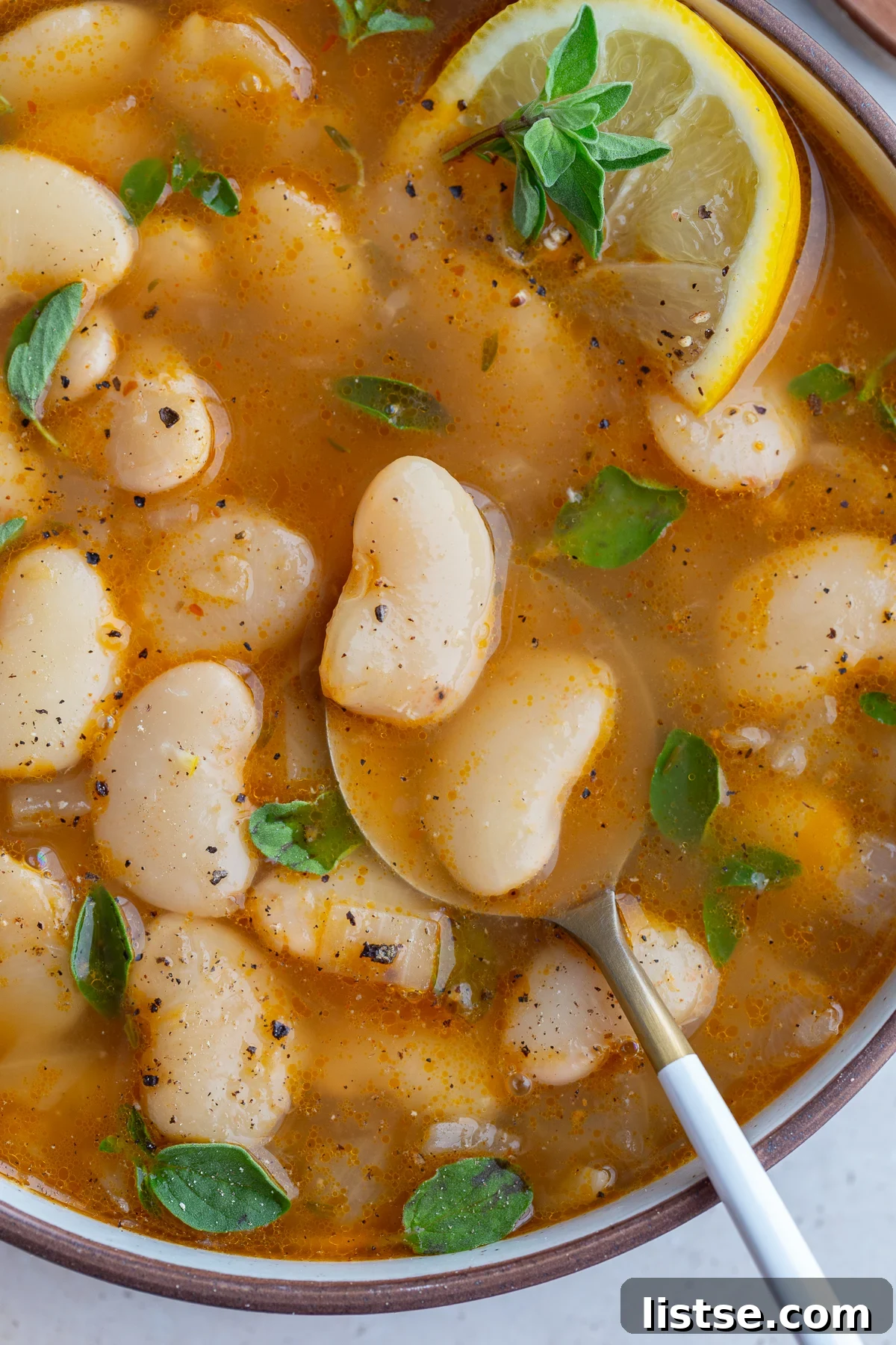 A close-up of brothy beans in a white bowl, garnished with a lemon wedge and a spoon, emphasizing the creamy texture of the beans and the rich broth.
