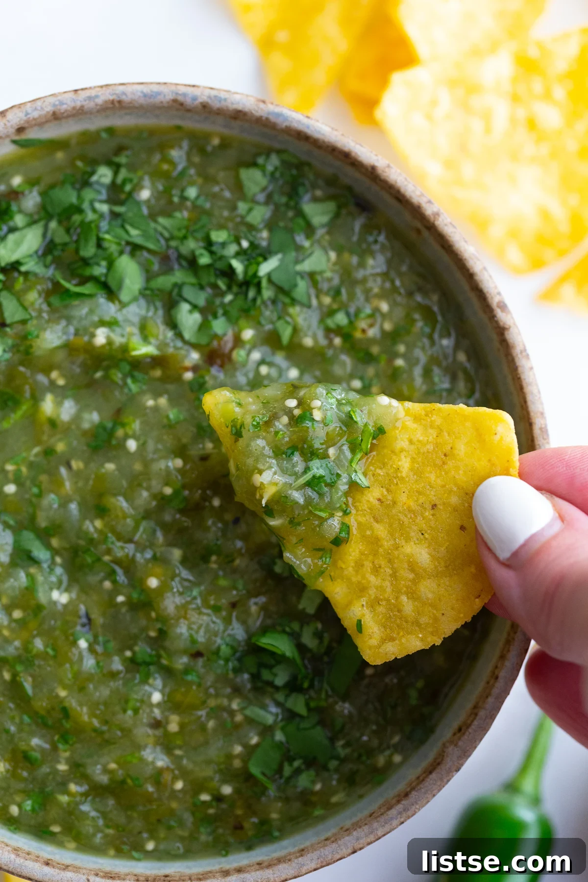 A hand holding a crispy tortilla chip dipping into a bowl of freshly made Roasted Salsa Verde, highlighting its appealing texture.