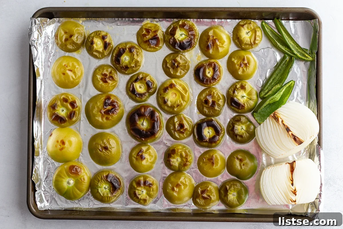 Halved tomatillos, peppers, and onion arranged on a foil-lined baking sheet after being roasted to a light char.
