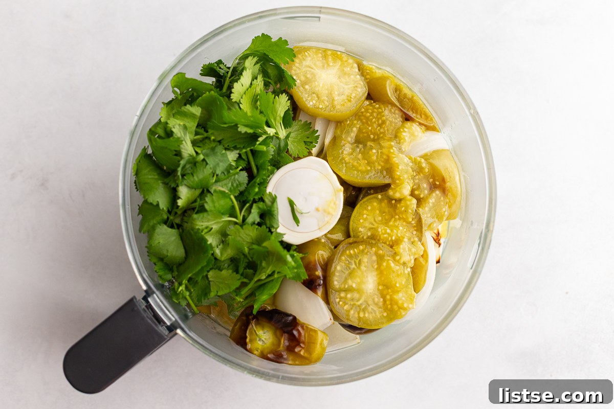 Roasted vegetables, including tomatillos, peppers, and onion, in a food processor with fresh cilantro.