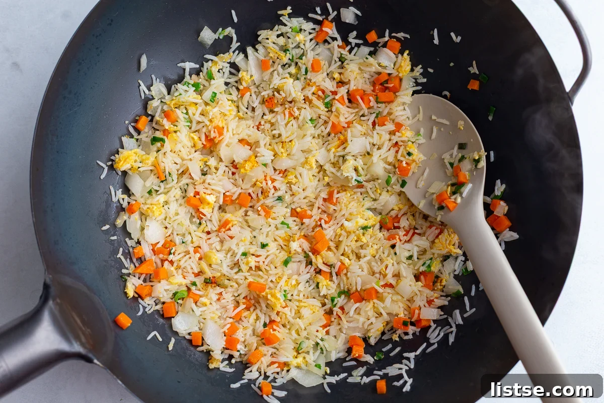 The initial stage of cooking fried rice in a large, hot wok, showing the process of stir-frying rice, onions, garlic, and carrots together, beginning to release their aromas.