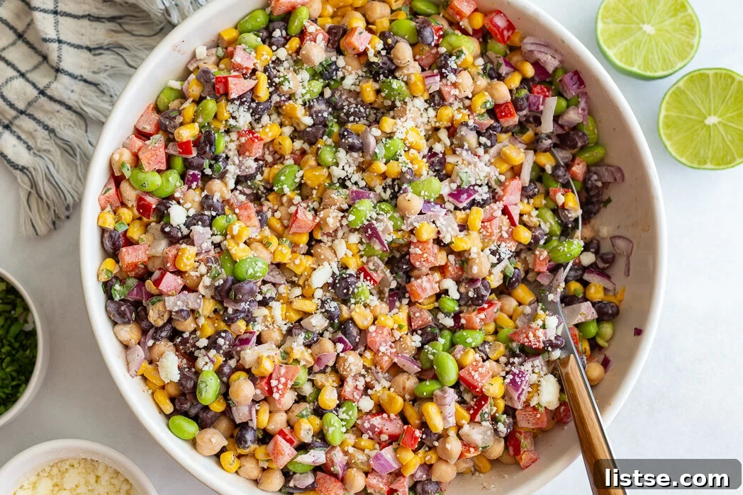 A large glass bowl filled with Mexican street corn bean salad, featuring chickpeas, black beans, corn, and red peppers, topped with crumbled cotija cheese.
