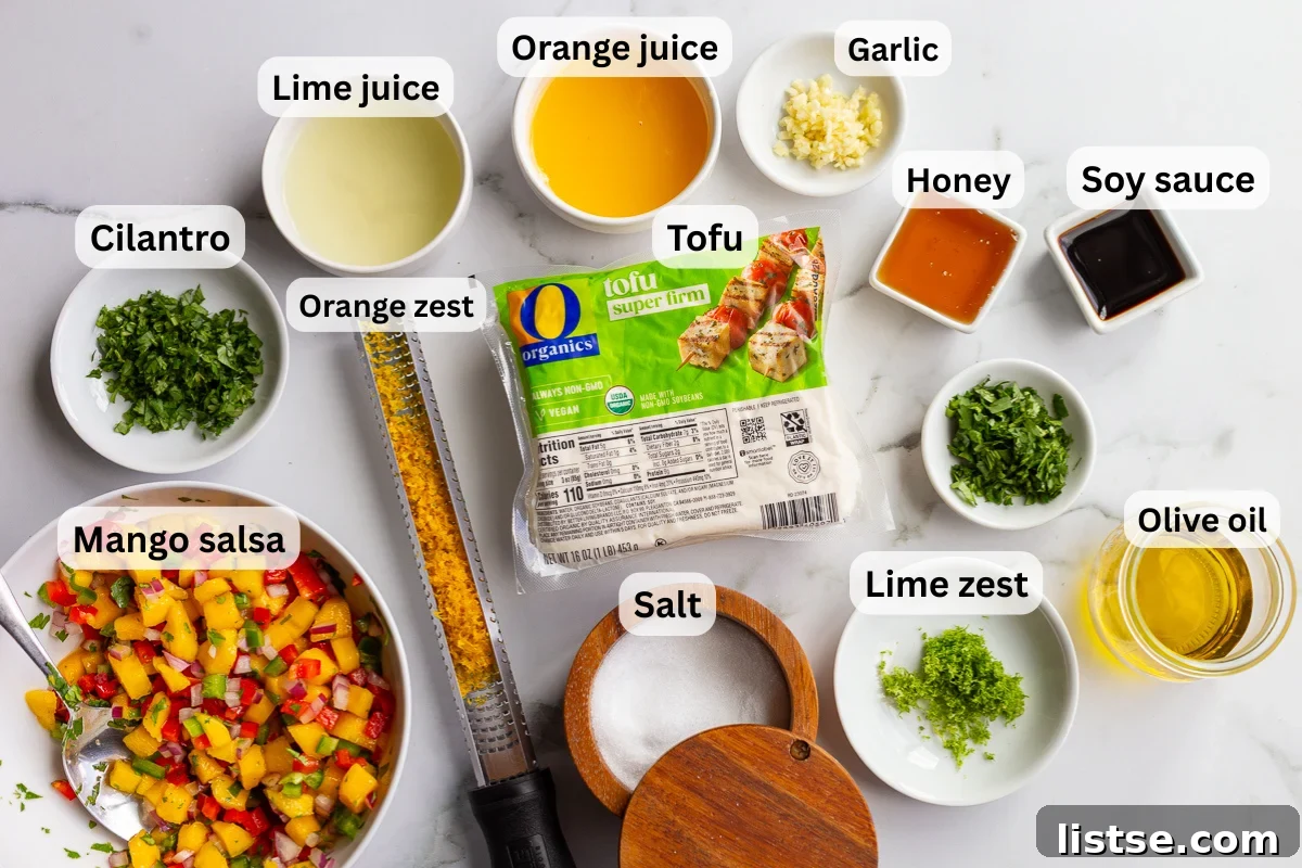 Cilantro-Lime Tofu with Fresh Mango Salsa 4 Overhead shot of cilantro lime tofu ingredients including super firm tofu, lime juice, lime zest, orange juice, orange zest, garlic, honey, soy sauce, cilantro, olive oil, salt, and a bowl of mango salsa, each labeled on a marble countertop.