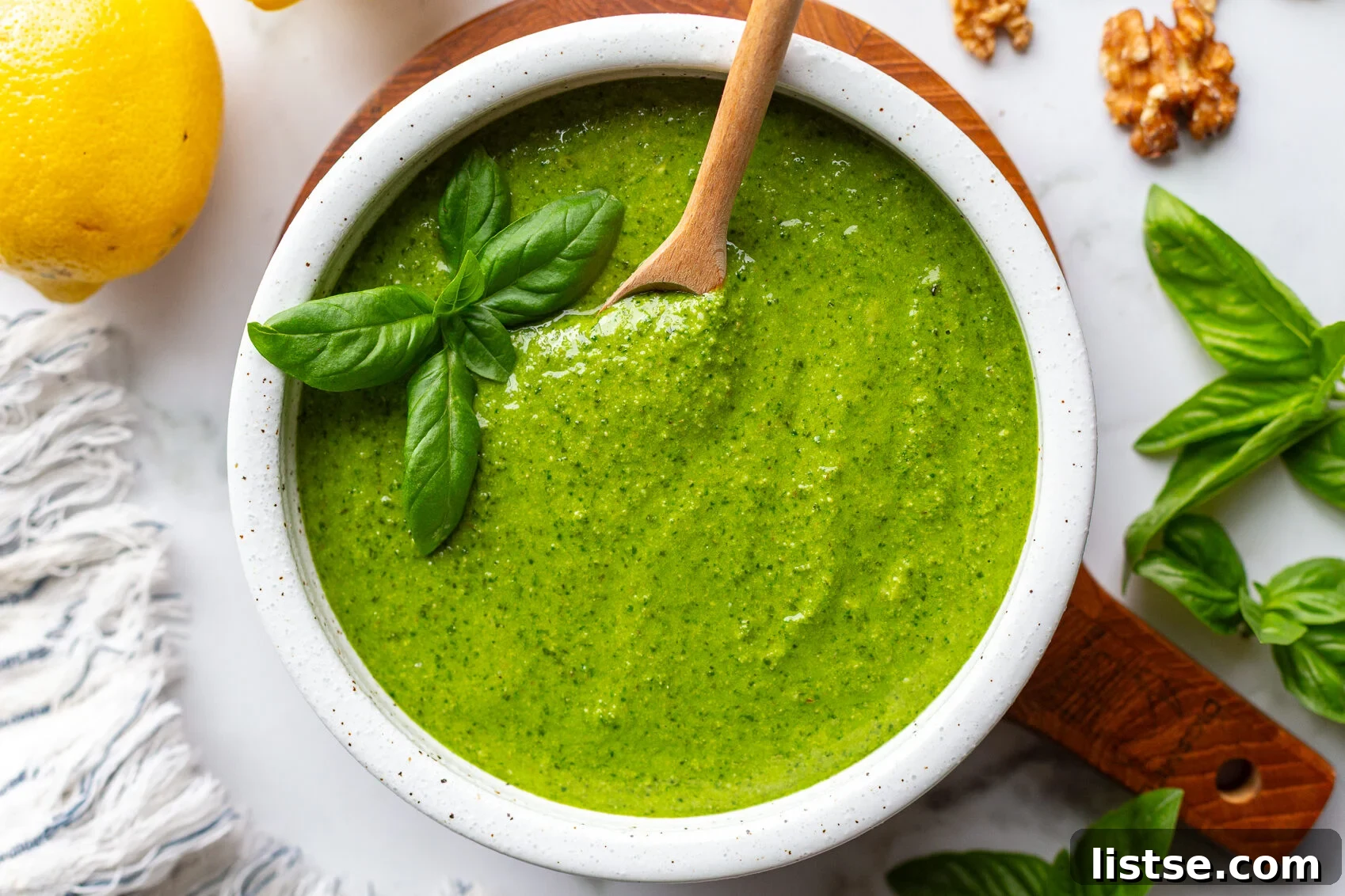 Homemade Walnut Pesto Recipe 10 Walnut pesto in a white bowl on a wooden board with a wooden spoon resting inside, surrounded by fresh basil, lemons, and walnuts